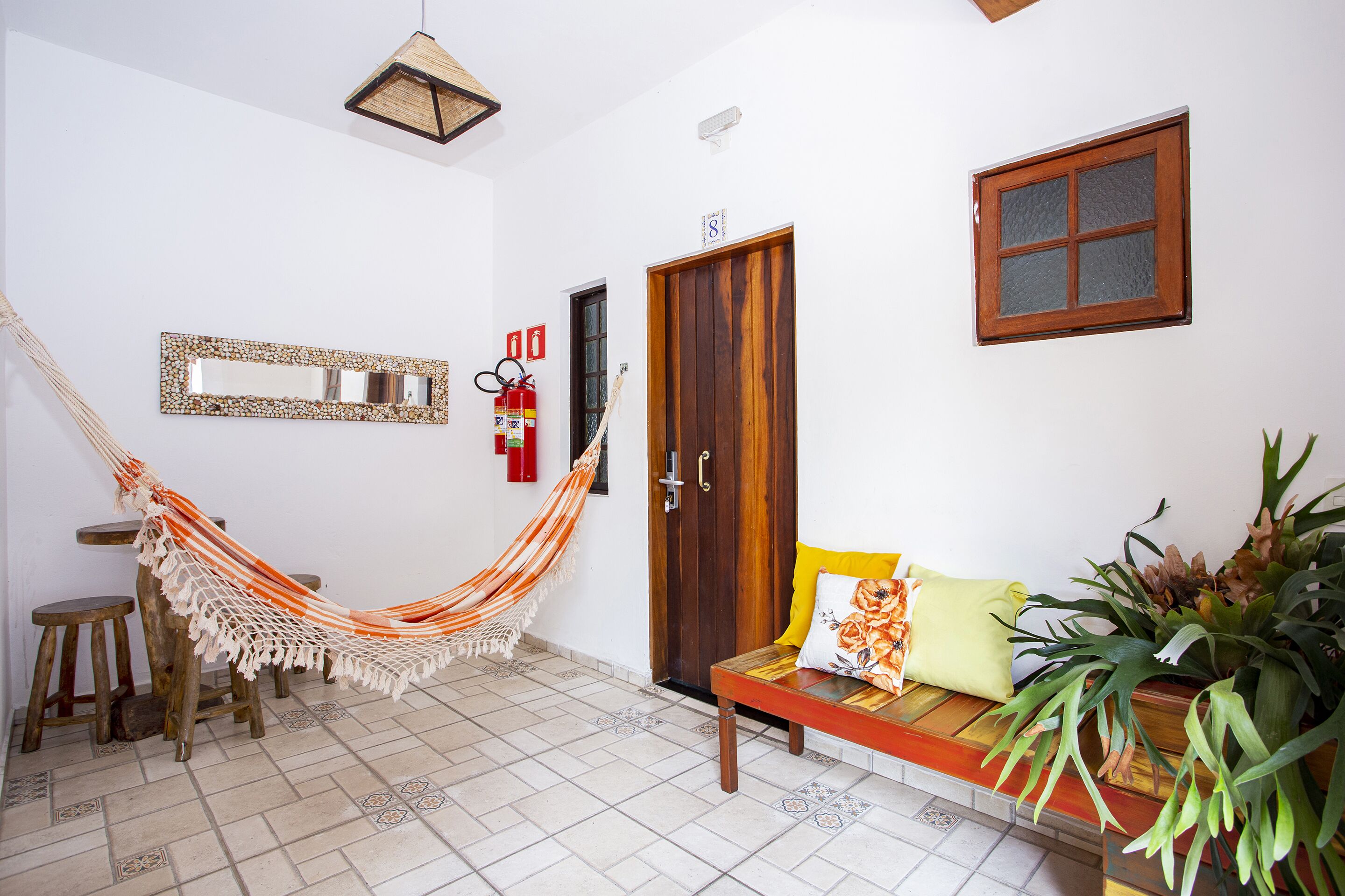 Una habitación con una hamaca, un banco de madera, un sofá con cojines, un espejo, una puerta y una ventana.