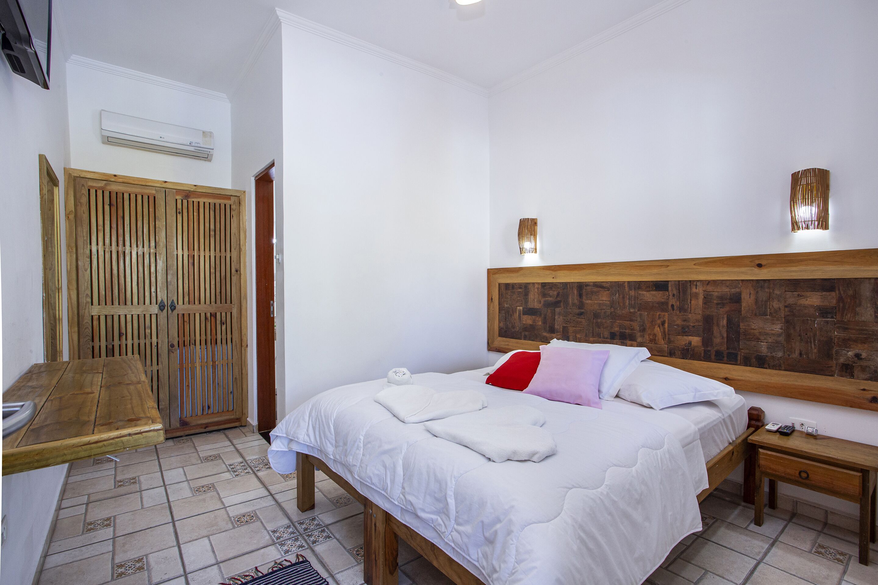 Ein Schlafzimmer mit einem Holzbett, weißer Bettwäsche, einem Nachttisch aus Holz, einem Beistelltisch, einem Holzschrank und einer Klimaanlage.