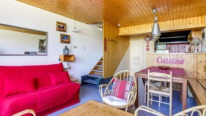 Une pièce chaleureuse avec un canapé rouge, des fauteuils en osier, une table en bois et un coin cuisine agrémenté d’une enseigne indiquant « Cuisine ».