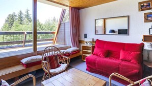 Une pièce chaleureuse avec un canapé rouge, des fauteuils en osier, une table en bois et une grande fenêtre donnant sur la forêt.