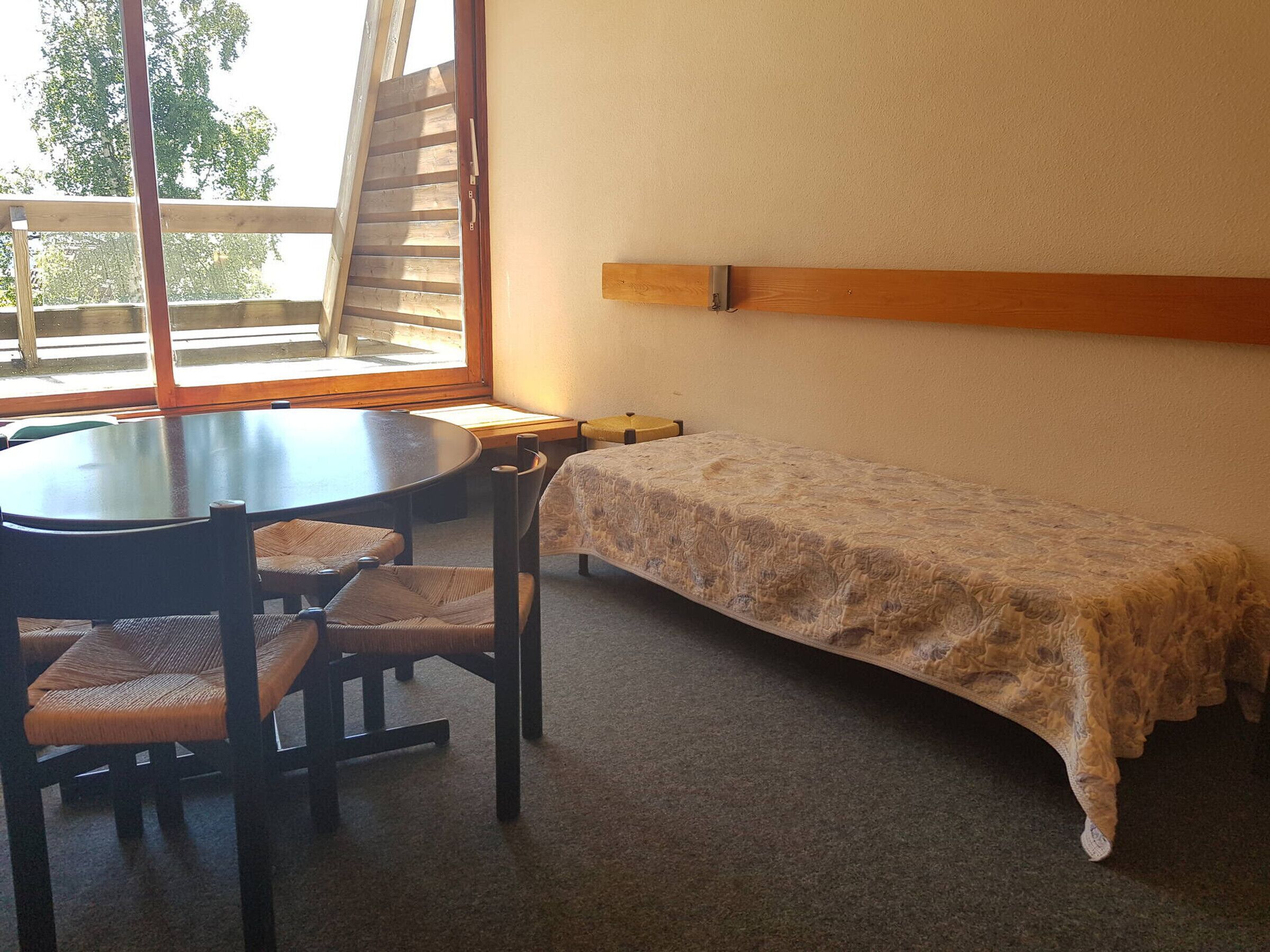 Une pièce avec une table ronde et quatre chaises, un petit lit recouvert d’une couverture à motifs, un banc en bois et une fenêtre donnant sur des arbres.