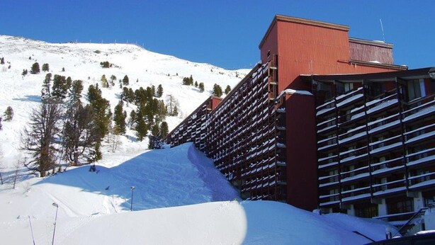 Un hôtel à la façade rouge, doté de nombreux balcons, avec une chaîne de montagnes enneigées en arrière-plan.