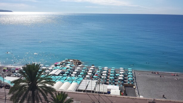 A beach with numerous umbrellas and sunbeds, a clear blue sea, and a distant shoreline. - 2 Bedroom Apartment 84m² Promenade des Anglais Last Floor sea View + Solarium (Nice)