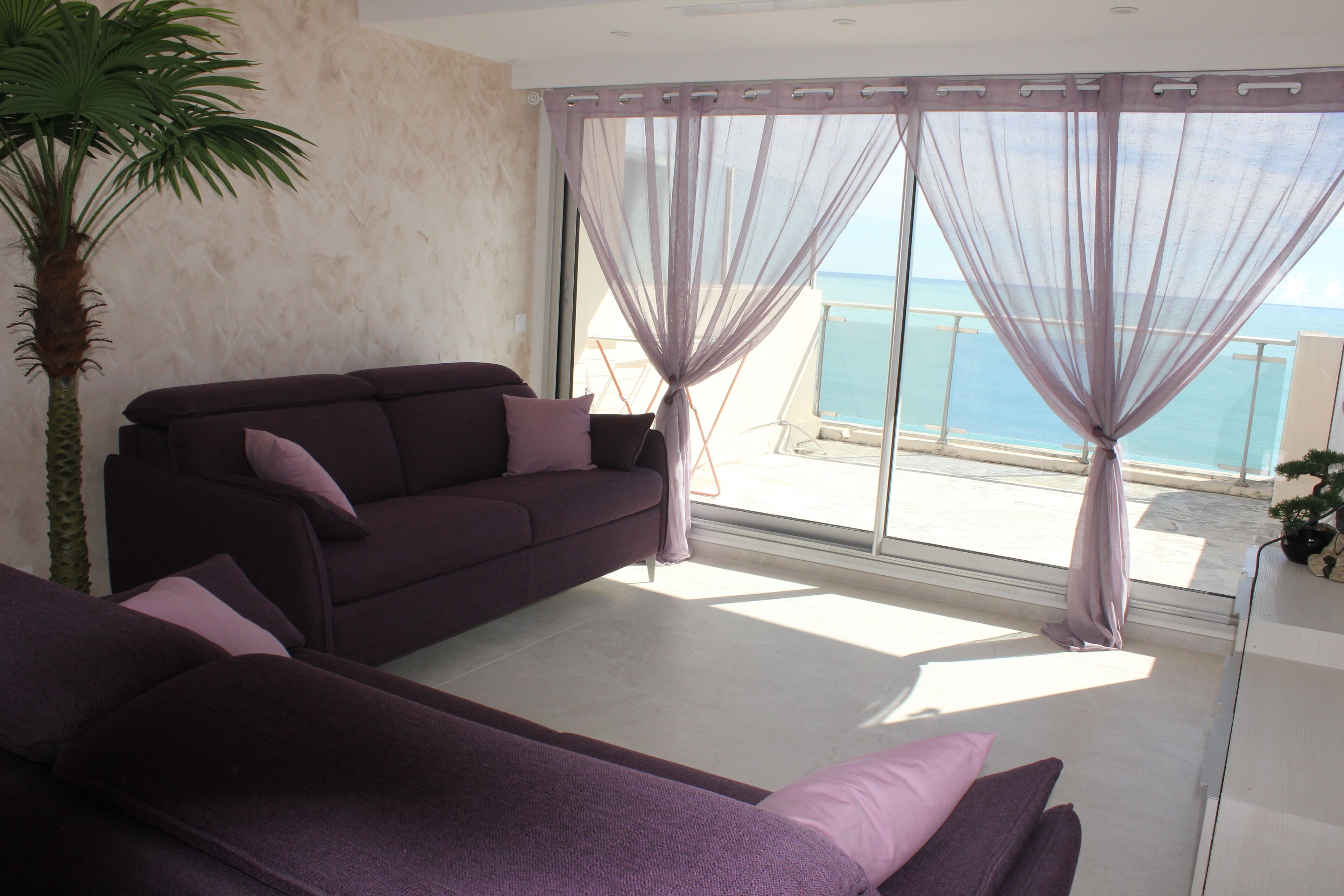 Un salón moderno con un sofá púrpura, un ventanal con cortinas translúcidas y vista al mar.