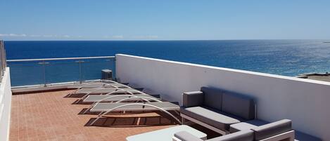 A rooftop terrace with a view of the sea, featuring a row of sun loungers and a white sofa.