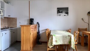 Pieni, käytännöllinen keittiö, jossa on puinen kaappi, kahvinkeitin ja imuri. Keittiössä on ruokapöytä tuoleineen sekä seinällä kehystetty taulu.