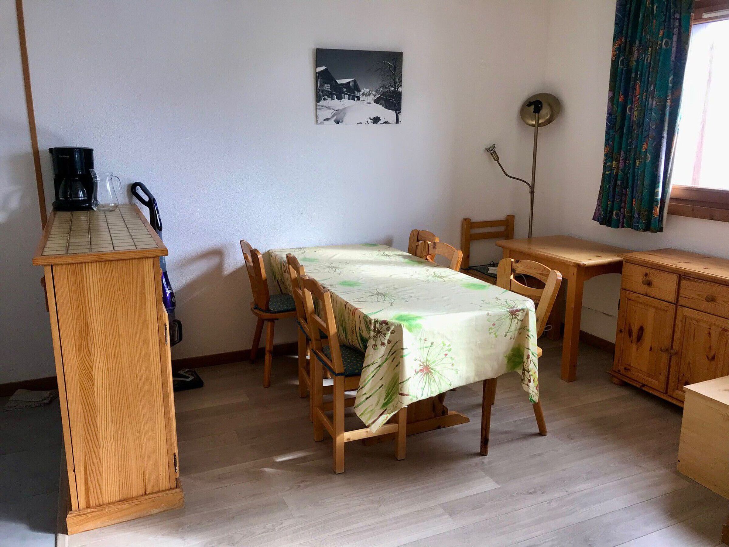 Un comedor con una mesa cubierta por un mantel floral, sillas de madera, un armario de madera y una cafetera.