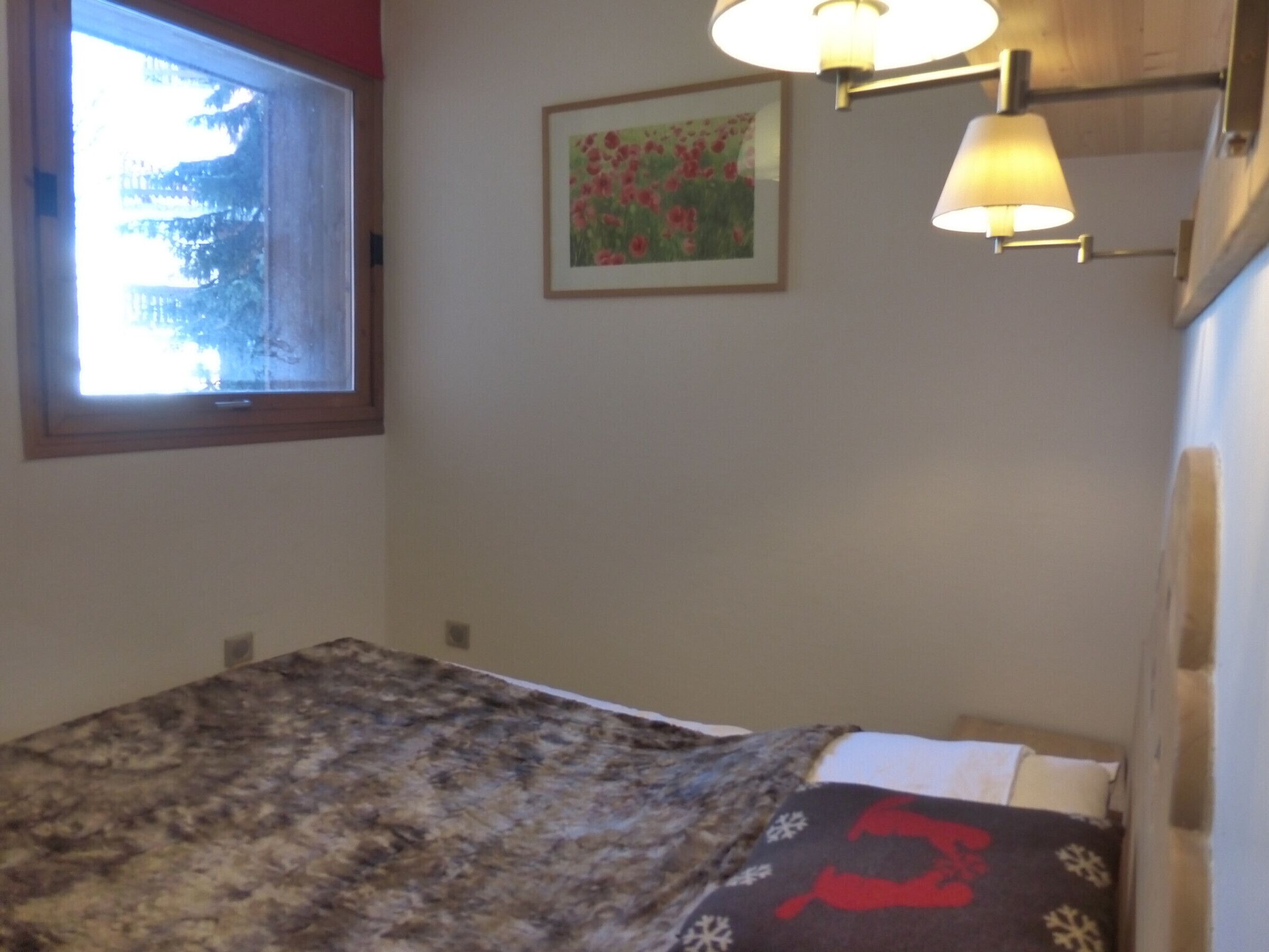 A bedroom with a bed, a window, a wall-mounted lamp, and a framed picture.