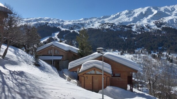 A wooden cabin with a snow-covered roof, set against a backdrop of snow-covered mountains and a clear blue sky.