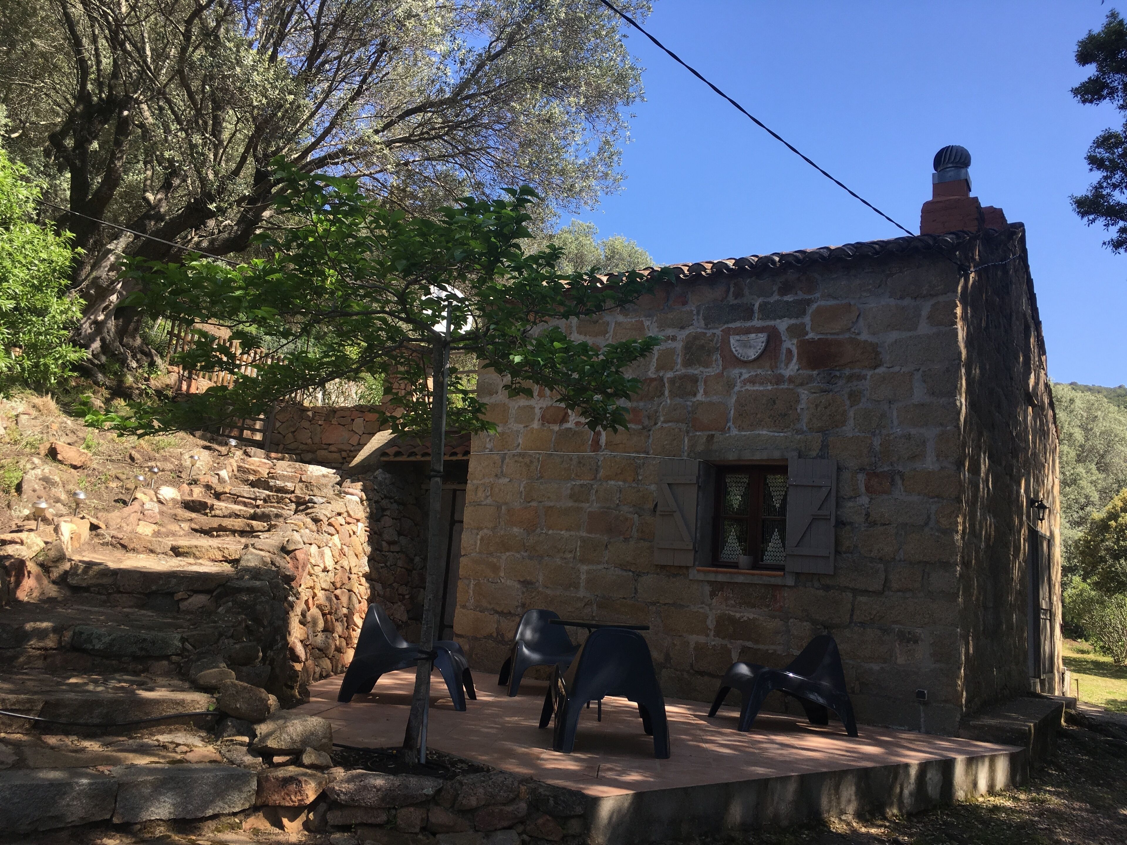 A rustic stone building with a tiled roof, a small window with shutters, and a stone staircase leading up.