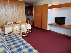 Ein kompakter Raum mit einem Bett, einem Esstisch mit Stühlen, einem Fernseher auf einem Regal und einem Kleiderschrank.