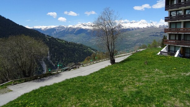 A modern building with a balcony, a paved path, and a mountainous landscape with snow-capped peaks in the background.
