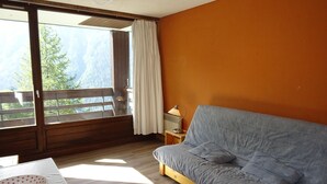 A room with a wooden floor, a grey sofa, a small table with a white tablecloth, and a window with a view of trees and mountains.