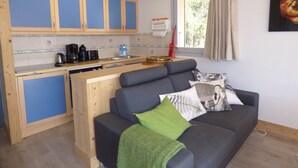 A compact kitchen with wooden cabinets and a built-in dishwasher. A grey sofa with a green throw blanket and patterned pillows is positioned in front of a window with a curtain. - Lovely apartment in Méribel center, close to slopes, for 5 persons (Méribel)