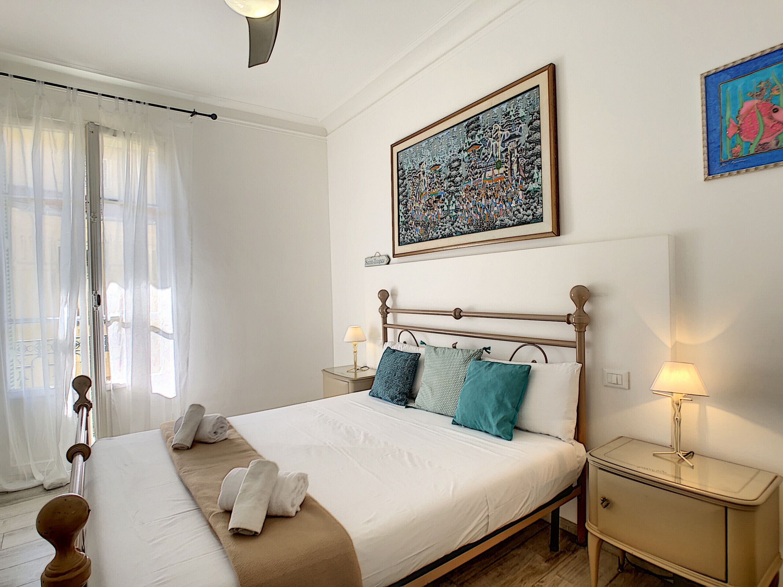 Un lit bien fait, avec du linge de lit blanc et un plaid marron, une table de chevet avec une lampe, et un tableau abstrait encadré accroché au mur.