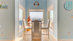 A dining area with a view of the beach, featuring a table with chairs, a decorative wall hanging, and a clock. - ☀️Dolphin Run: Oceanfront w/ Private Pool-Large Beachfront Deck-Pet Friendly☀️ (Kure Beach)