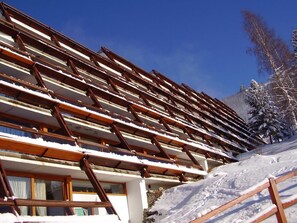 Un immeuble à plusieurs étages, doté d’un toit en pente, qui ressemble probablement à un hôtel ou à un complexe touristique, avec une vue sur une montagne enneigée en arrière-plan.