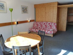 Een knus vertrek met een ronde houten tafel, stoelen, een houten bank met kussens met hartmotief en een groene vloerlamp.