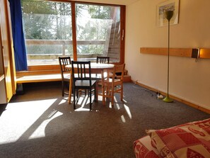Een kamer met een houten tafel en stoelen, een raam met uitzicht op bomen, een vloerlamp en een bed met een geborduurde deken.