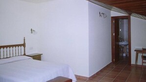 Quarto com cama, mesa de cabeceira, porta de madeira e banheiro visível através de uma porta aberta.