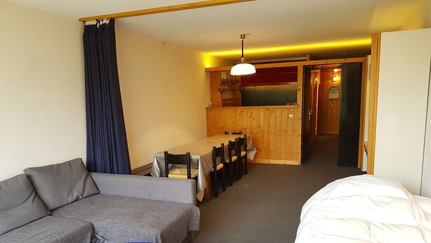 Une chambre d’hôtel comprenant un canapé, une petite table bleue, un lit, un coin repas avec une table et des chaises, et un coin cuisine avec un comptoir et des luminaires suspendus.