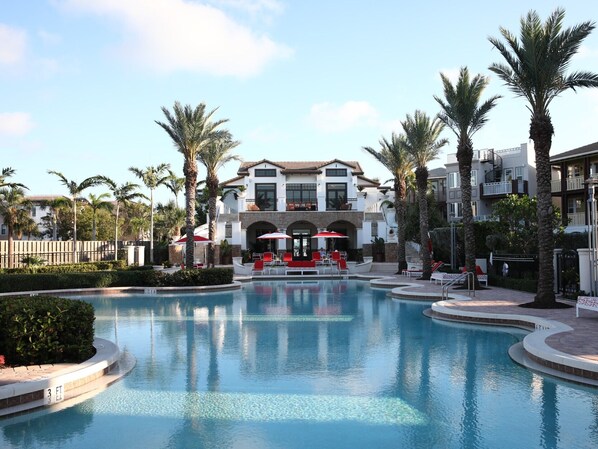 A swimming pool with lounge chairs and umbrellas, surrounded by palm trees and a building with balconies.