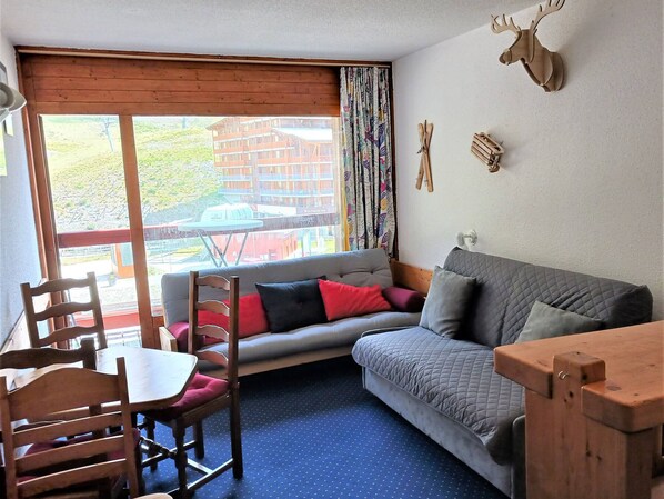 A cozy room with a wooden table, a grey sofa with red and black cushions, a blue carpet, and a large window with a view of a building and greenery outside. - Ski-in/ski-out studio for 4 at Arc 2000 (Les Arcs 2000)