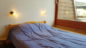 Un lit simple avec une tête de lit et un pied de lit en bois, une couverture grise et deux lampes fixées au mur.