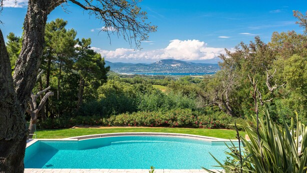 A clear view of a swimming pool surrounded by lush greenery and trees, with a distant mountain range in the background.