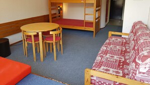 Un lit superposé avec un matelas rouge, une table en bois avec quatre chaises, un canapé rouge et une petite poubelle noire.