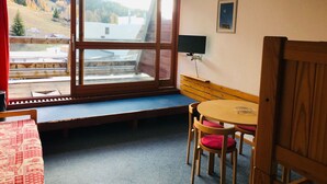 Une pièce dotée d’une grande fenêtre, d’un banc en bois, d’une petite table avec des chaises et d’un téléviseur fixé au mur.