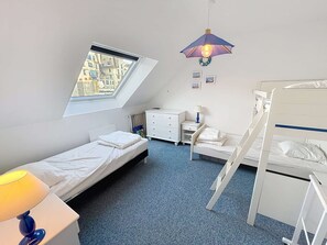 Et soveværelse med køjeseng, enkeltseng, skrivebord med lampe, kommode og vindue med udsigt til bygninger.