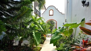 Un sendero estrecho que lleva a un edificio de fachada blanca y techo de tejas rojas, rodeado de exuberante vegetación tropical.