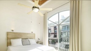 Un dormitorio con cama, ventilador de techo, mesita de noche y una ventana grande con cortinas.