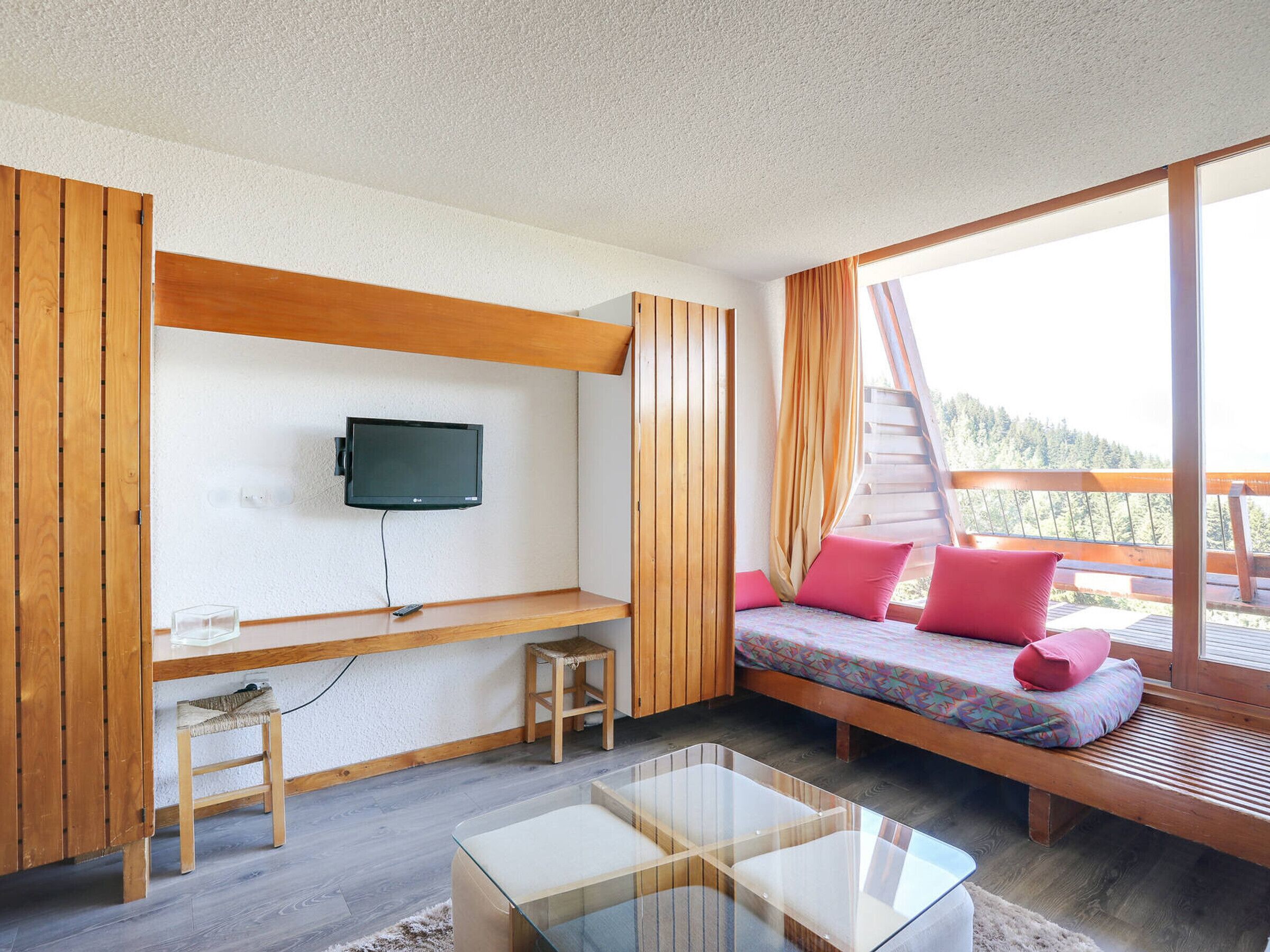 A cozy room with a bed, a TV mounted on the wall, a small table, and a window with a view of the outdoors.