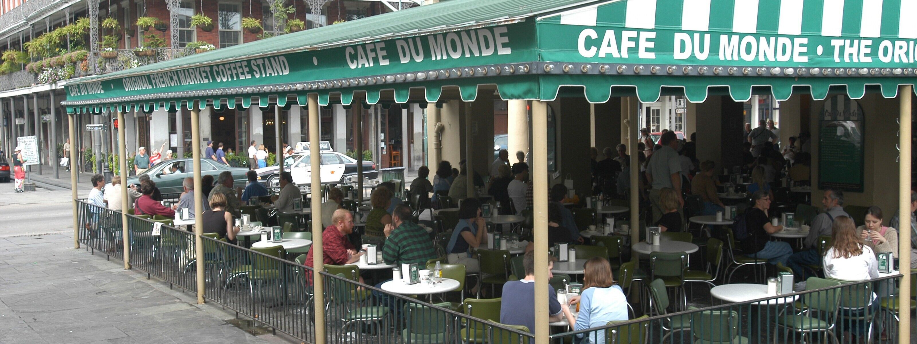 An outdoor café with tables and chairs, people dining, and a green awning with signage.