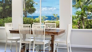 Un comedor con mesa de madera y sillas blancas, ventanas amplias con vista al océano y follaje.