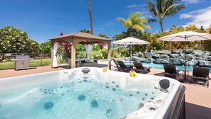 Un jacuzzi con sillones de descanso y sombrillas, un pabellón y una piscina con cascada de fondo.