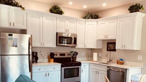 A modern kitchen with white cabinets, a stainless steel refrigerator, a microwave, and an oven. There is a dining area with a wooden table and chairs.