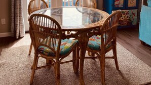 A dining area with a glass-top table surrounded by wicker chairs, a blue cabinet with colorful artwork, and a view of a balcony with a railing.