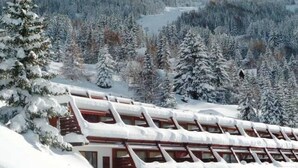 Una cabaña cubierta de nieve con techo inclinado y varias ventanas, rodeada de un bosque nevado.