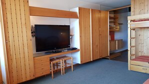 Una habitación con un televisor de pantalla plana grande sobre un soporte de madera, un escritorio de madera pequeño con una silla, y una cama individual con un edredón rojo.