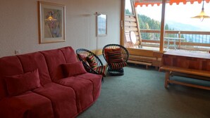 Un salón con un sofá rojo, sillas de mimbre, una mesa de madera y un ventanal con vistas a las montañas.