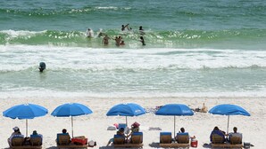 Une plage où l’on voit des gens se baigner et d’autres, assis sous des parasols bleus, se détendre sur le sable.