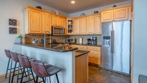 A kitchen with wooden cabinets, a stainless steel refrigerator, a microwave, a stove, and a bar stool area.