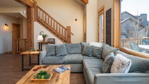 A modern living room with a sectional sofa, a wooden coffee table with a magazine and plants, a staircase with wooden railings, and a large window with a view of buildings.