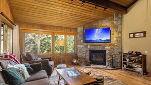 A cozy living room with a stone fireplace, a large TV mounted on the wall, and a comfortable seating area with a sofa and chairs. - Walk to Ski & Village, Views Condo, Pool & Spa 315 (Mammoth Lakes)