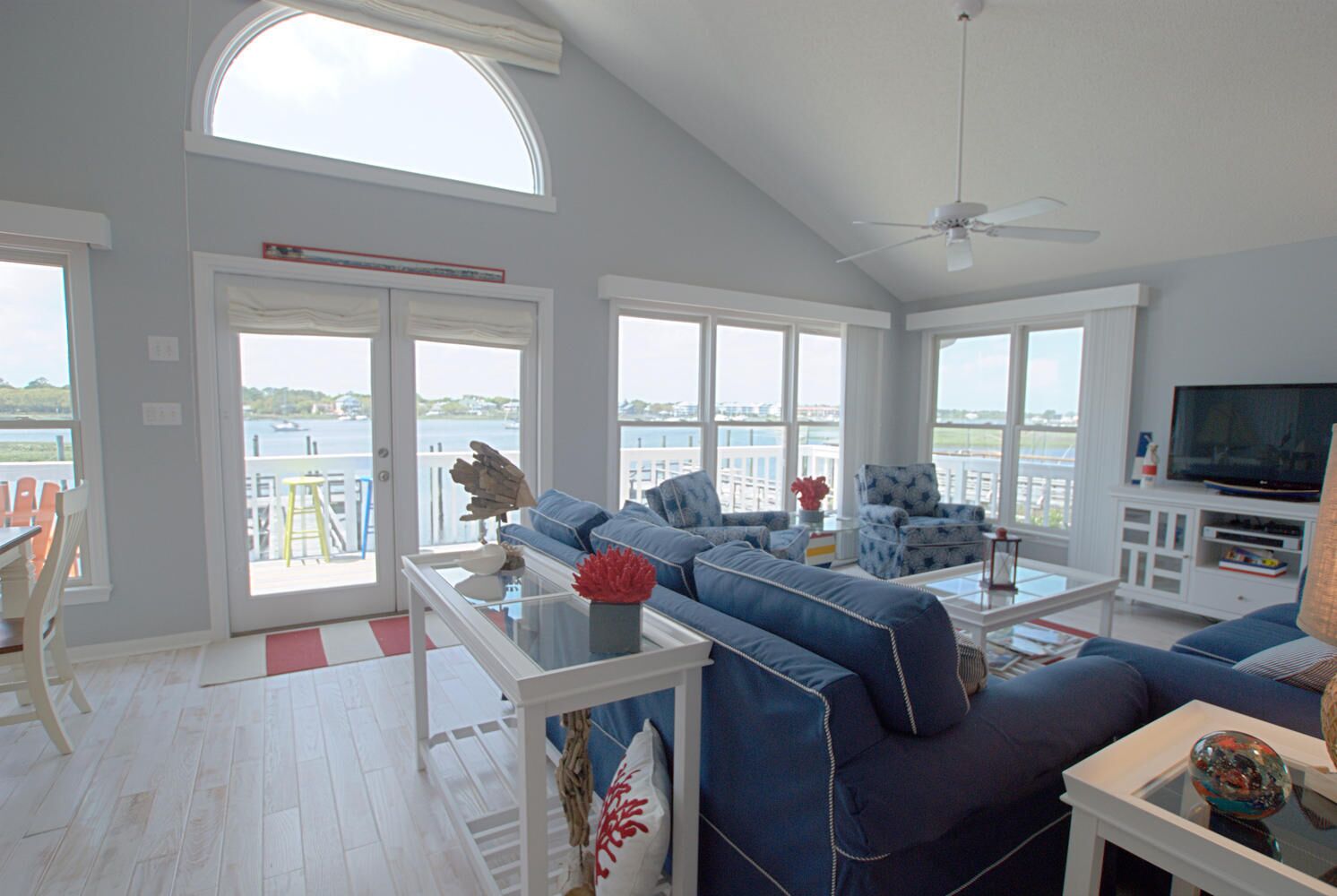 A spacious living room with a large window overlooking a body of water, a ceiling fan, and a television on a white cabinet.