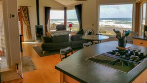 A modern living room with a fireplace, a sofa, a dining table, and a kitchen area with a stove. Large windows offer a view of the ocean.