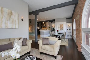 Amplia sala de estar con un sofá color beige, una mesa de centro y una planta de orquídea blanca. Se ve una cocina con electrodomésticos modernos y un comedor con mesa y sillas al fondo.